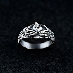 This sterling silver ring combines the gothic allure of skeleton hands with the timeless symbolism of the Claddagh ring, representing love, loyalty, and friendship. The intricate design features two skeleton hands holding a heart, crowned by a subtle flame motif, making it an ideal piece for those who embrace dark, edgy aesthetics with deep meaning. Details:     Material: 925 Sterling Silver     Finish: Oxidized highlighting its refined and timeless appeal. Our pieces are crafted with love and m Gothic Skull Open Ring Gift, Adjustable Gothic Skull Ring, Hand Cast Skull Ring For Halloween Gift, Gothic Metal Skull Ring For Promise, Gothic Metal Skull Ring As Promise Ring, Gothic Silver Sterling Silver Skull Ring, Symbolic Skull Rings For Halloween, Halloween Skull Rings Symbolic Style, Vintage Sterling Silver Rings For Halloween