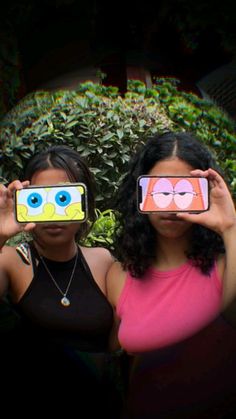 two women are holding up their cell phones to show the same faces as they stand in front of some bushes