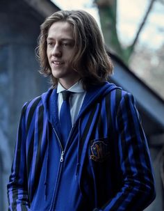 a man with long hair wearing a blue striped jacket and tie, standing in front of a building