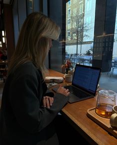 a woman sitting at a table with a laptop computer in front of her, looking out the window