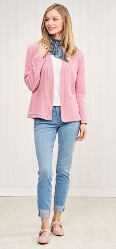 a woman standing in front of a white wall wearing jeans and a pink cardigan