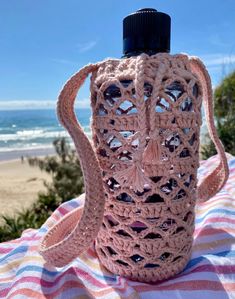 a pink crocheted water bottle sitting on top of a blanket next to the ocean