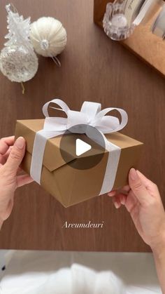 someone is holding a brown box with a white ribbon and bow on it, while sitting at a table