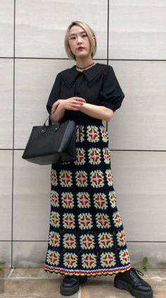 a woman standing in front of a wall with her hand on her hip, wearing a black shirt and multicolored skirt