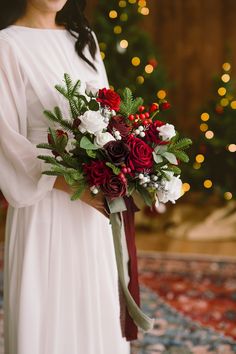 a woman in a white dress holding a red and white bouquet with greenery on it