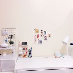 a white desk topped with lots of clutter next to a lamp and pictures on the wall