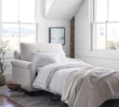 a bed with white sheets and pillows in a room next to two large windows on the wall