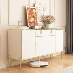 a white cabinet with gold legs and flowers on top in front of a large window