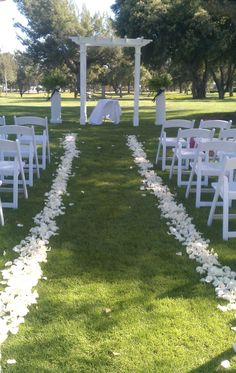 an outdoor ceremony setup with white chairs and petals on the grass, in front of a pergolated arch