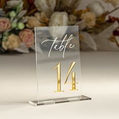 a clear acrylic table number holder with gold lettering and flowers in the background