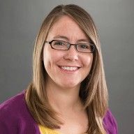 a woman wearing glasses and a purple jacket smiles at the camera while standing in front of a gray background