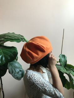 a woman wearing an orange hat and holding a plant in front of her face while standing next to a white wall