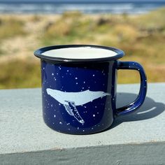 a blue coffee mug with a white whale painted on it's side and the ocean in the background