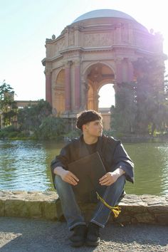 a man sitting on the ground in front of a pond with a building behind him