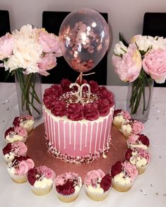 a pink and white cake surrounded by cupcakes on a table next to flowers