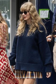 a woman in a blue sweater and plaid skirt is walking down the street with her hand on her hip