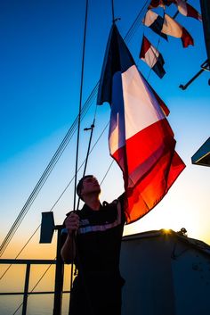 a man holding a red, white and blue flag on top of a boat at sunset