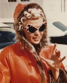 a woman in an orange coat and sunglasses making the peace sign with her hand while standing next to a car