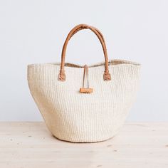 Rural Kamba women in Kenya hand-weave these beautiful handbags from tough sisal fibers. Sisal grows well in the semi-arid climate of eastern Kenya, and its spiky leaves contain long fibers that can be extracted for weaving.  approximate size: 10" diameter x 10" tall (without handle), 17" tall (with handle) leather hand Eco-friendly Woven Straw Travel Bag, Eco-friendly Woven Straw Bag For Travel, Eco-friendly Weaved Straw Bag For Travel, Natural Straw Bag Fair Trade, Eco-friendly Straw Travel Bag With Weaving, Natural Straw Fair Trade Bag, Eco-friendly Fair Trade Natural Beach Bag, Artisan Beige Straw Bag For Market, Natural Weaved Straw Bag For Travel