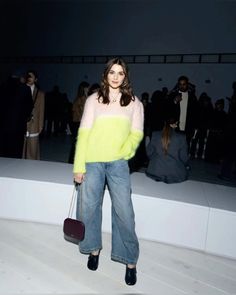 a woman standing in front of a group of people wearing jeans and sweaters, holding a handbag