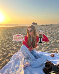 a woman sitting on top of a beach next to a bottle of wine and cake