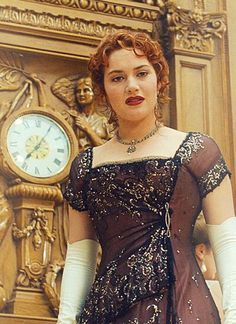 a woman in a brown dress and white gloves standing next to a wall with a clock