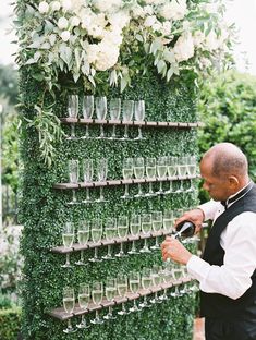 a man standing next to a tall green wall filled with glasses and flowers on top of it