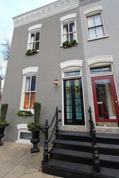 a grey house with red door and black steps leading up to the front door, surrounded by potted plants