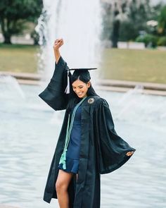 a woman wearing a graduation gown and holding her arms up in the air while standing next to a fountain