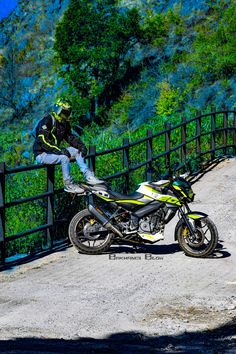 a man riding on the back of a yellow and black motorcycle next to a lush green hillside