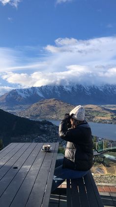 a woman sitting on top of a wooden bench next to a lake and snow covered mountains