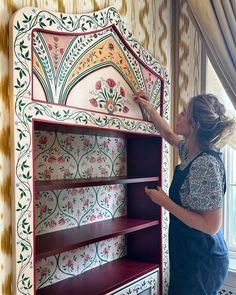 a woman in an apron is painting a wallpapered bookcase with floral designs