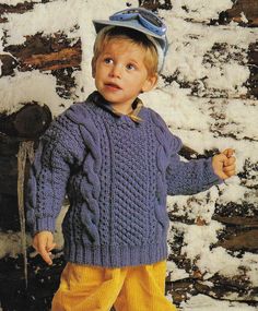 a young boy wearing a blue sweater and yellow pants standing in front of snow covered rocks