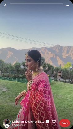 a woman in a pink sari and gold jewelry standing on grass with mountains in the background