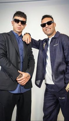 two men standing next to each other in front of a white wall wearing suits and sunglasses