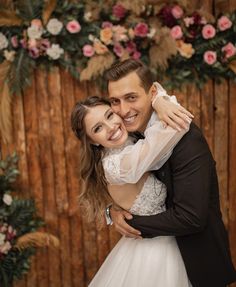 a bride and groom hugging each other in front of a wooden wall decorated with flowers