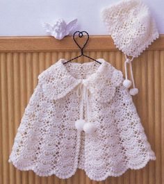 a white crocheted baby coat and hat hanging on a wooden wall next to a bird ornament