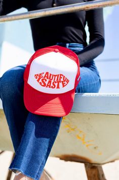 a woman sitting on a bench wearing a red and white trucker hat with the words beautiful sasy printed on it