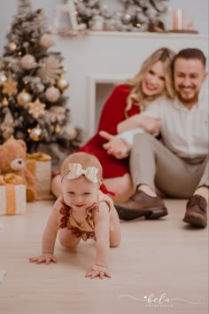 a baby crawls on the floor in front of a christmas tree with her parents