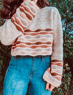 a woman standing in front of some bushes wearing a sweater and jeans with her hands on her hips