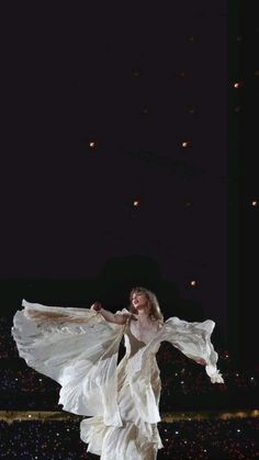 a woman in white dress standing on stage with her arms spread out and lights behind her