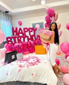 a woman in white dress sitting on bed with pink balloons and presents for her birthday