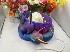 a purple bowl with yarn and knitting needles in it next to some flowers on a table