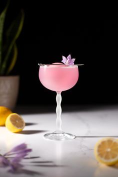 a pink cocktail garnished with an orchid sits on a table next to lemons