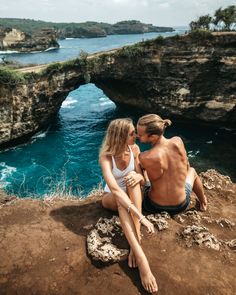 two people are sitting on the edge of a cliff by the ocean and looking at each other