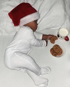 a baby in a santa hat eating cookies and milk on a bed with white sheets