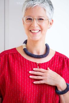 a woman wearing glasses and a red top smiles at the camera with her hand on her chest