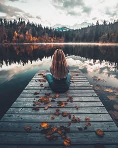 a woman sitting on a dock in front of a lake with trees and leaves around her