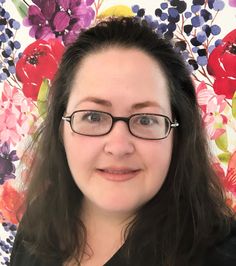 a woman wearing glasses standing in front of a flowered wall with flowers on it