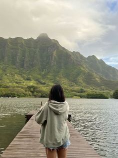 a woman standing on a dock looking at the water with mountains in the background,